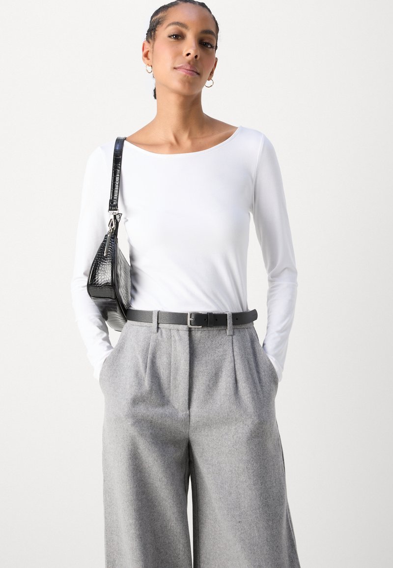 Top bianco a maniche lunghe abbinato a pantaloni grigi a vita alta con pieghe. Gli accessori includono una borsa nera in tessuto texturizzato e una cintura nera sottile.