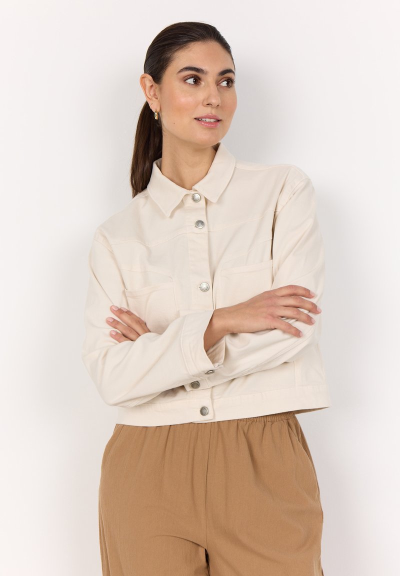 Veste courte couleur crème, en coton avec un devant boutonné, deux poches poitrine et un col classique. Texture lisse avec des boutons en argent.