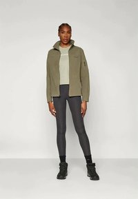 Olijf groene jas met een hoge kraag, gedragen over een lichtgrijze shirt en zwarte legging, aangevuld met zwarte wandelschoenen.