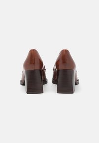 Zapatos de tacón de cuero marrón con tacón cuadrado, con textura suave y costuras mínimas. La vista trasera resalta la forma del tacón y el contraste de material.