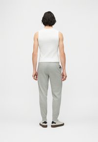 Άτομο με σκουρόχρωμα σγουρά μαλλιά στέκεται γυρισμένο προς τα πίσω, φορώντας λευκό αμάνικο τοπ, γκρι φόρμα και λευκά αθλητικά παπούτσια με μαύρες λεπτομέρειες.