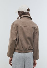 Braune Fleecejacke mit hohem Kragen, elastischem Saum und langen Ärmeln, kombiniert mit einer beigen Mütze. Graue Jogginghose vervollständigt das Outfit.