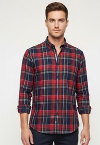 Camisa de cuadros en rojo y azul marino con cuello abotonado, mangas largas remangadas y logo en el bolsillo izquierdo del pecho. La tela parece texturizada.