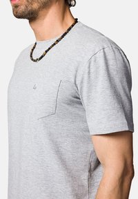 Szara koszulka z krótkim rękawem z miękkiego materiału z małym detalem kieszeniowym. Naszyjnik z koralików z czarnymi i brązowymi koralikami noszony na szyi.