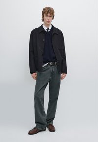 Sort yderjakke, marineblå striktrøje, hvid skjorte, grå jeans, brune sko. Simpelt design med knapper og diskrete teksturer.