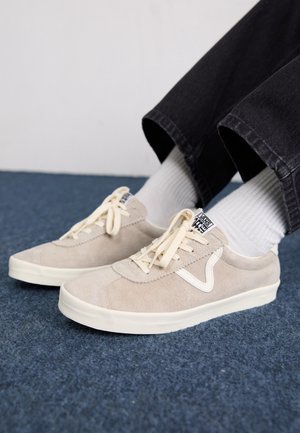 Personne portant des baskets Vans en daim beige avec des semelles blanches, des chaussettes blanches et un pantalon noir, assise sur un sol tapissé bleu.