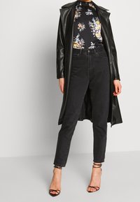 Un long manteau en similicuir noir porté par-dessus un blouse florale noire et un jean taille haute noir. Associé à des sandales à brides noires.