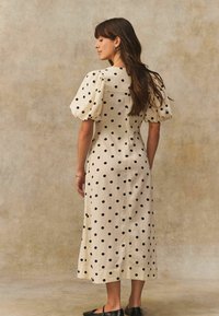 Kleid mit Cremefarbenen Polka-Dots, Puffärmeln, mittellanger Silhouette, verborgenem Reißverschluss im Rücken und glattem Stoff. Schwarze Akzente auf hellem Hintergrund.