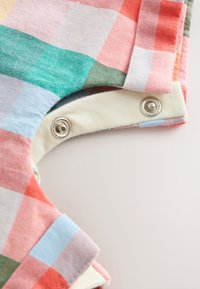 Tissu à carreaux multicolores avec boutons-pression métalliques argentés sur une doublure couleur crème, montrant un bord de vêtement et un détail d’attache.