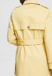 Trench-coat jaune avec une taille ceinturée, une grande rabat au dos et des détails à boutons. Tissu lisse et léger avec des manches longues et une coupe ajustée.