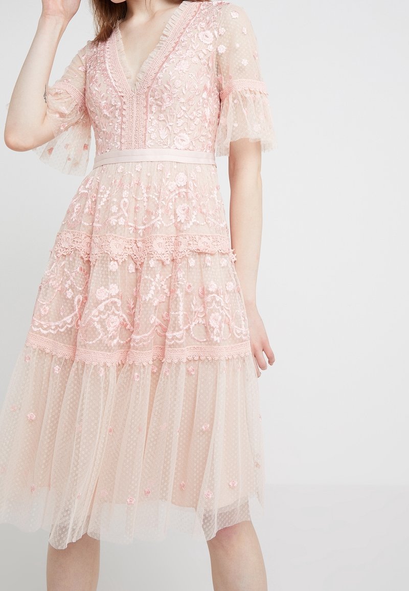 Femme en robe en dentelle rose transparente longueur genoux, brodée de motifs floraux, manches courtes à volants et col en V profond sur un fond uni.