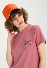 T-shirt a righe rosse e bianche con una tasca a forma di cuore ricamata in nero, abbinata a un cappellino arancione brillante. Capelli ricci in vista.