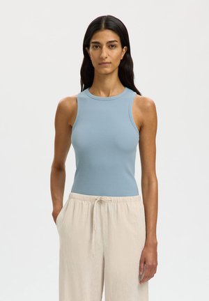 Kobieta z długimi ciemnymi włosami, ubrana w bezrękawnik w jasnoniebieskim kolorze z prążkowanego materiału oraz beżowe spodnie z wiązaniem w pasie, stojąca z rękami w kieszeniach.