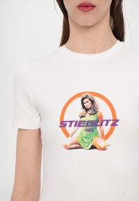 Hvit bomulls-T-skjorte med en fargerik grafisk utskrift av en sittende kvinne i en grønn kjole, med "STIEGLITZ 1988" i lilla og oransje aksenter.