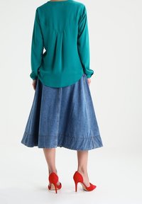 Blusa de manga larga en color azul verdoso con detalle de pliegues, combinada con una falda midi de mezclilla y unos zapatos de tacón rojo.