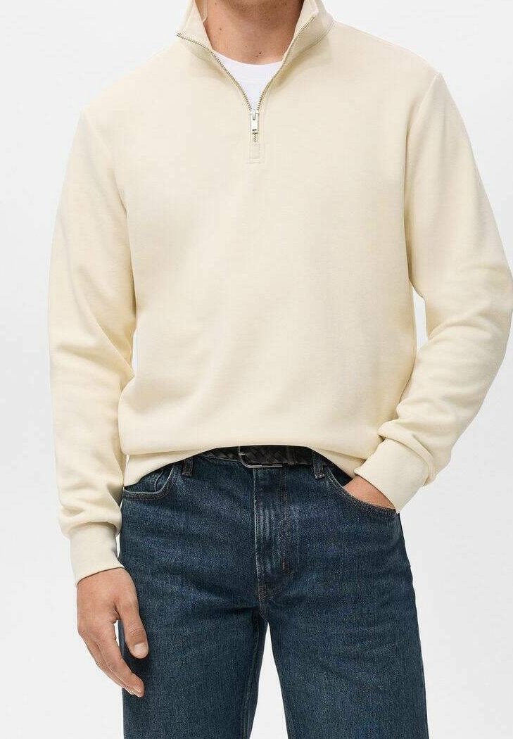 Homme portant un pull crème à fermeture éclair au quart et un jean bleu foncé, une main dans la poche et l'autre détendue sur le côté.