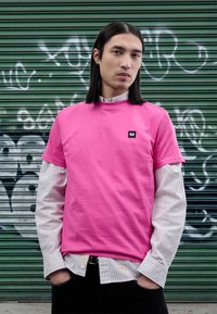 Camiseta de algodón rosa con mangas cortas, superpuesta sobre una camiseta de manga larga a rayas blancas y azul marino; cuenta con un pequeño parche de logo negro.