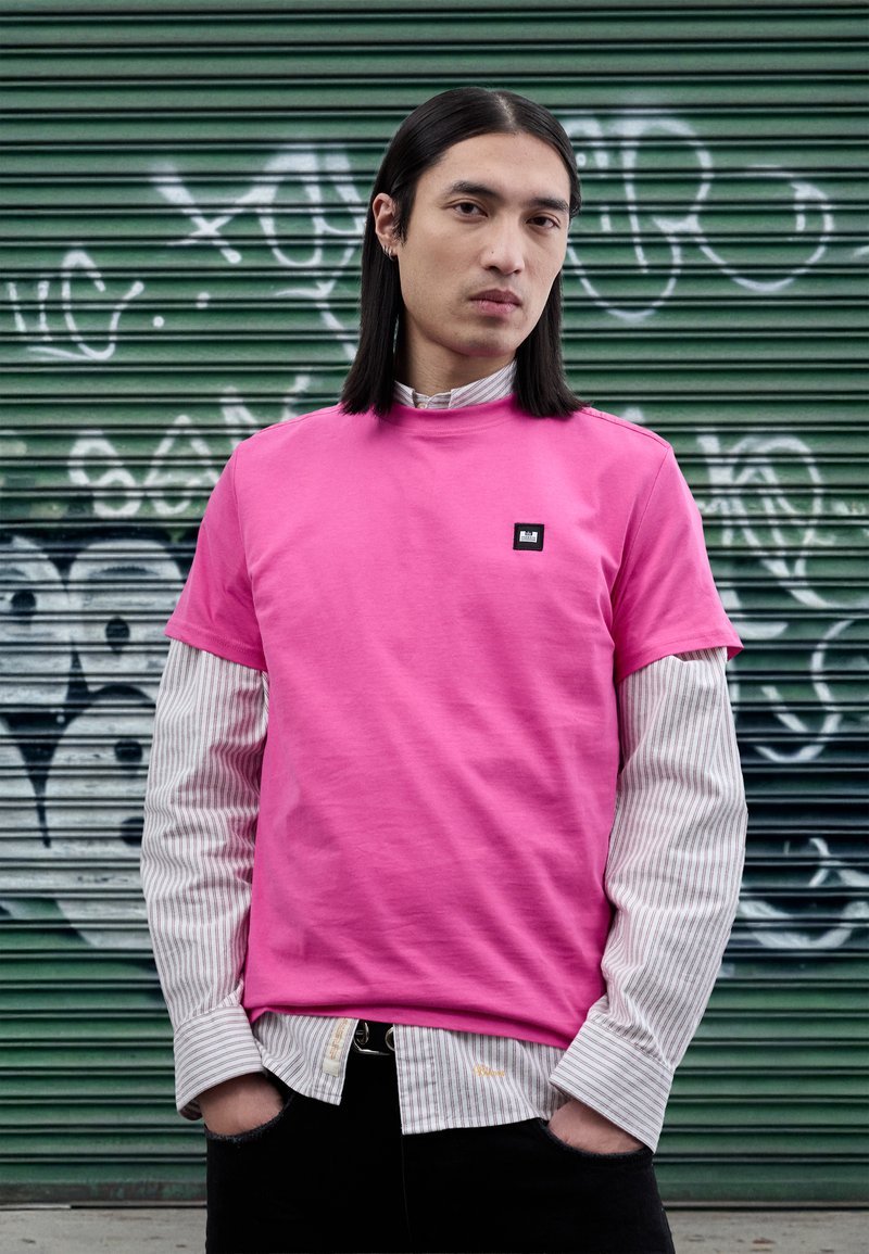 Camiseta de algodón rosa con mangas cortas, superpuesta sobre una camiseta de manga larga a rayas blancas y azul marino; cuenta con un pequeño parche de logo negro.