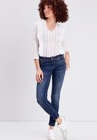 Blusa blanca de encaje con acentos de volantes, combinada con jeans ajustados de color azul oscuro. La modelo lleva zapatillas blancas. El conjunto tiene un ajuste casual.