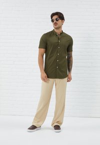 Short-sleeve olive green shirt with a button-down collar and chest pocket, paired with light beige trousers and dark brown loafers.