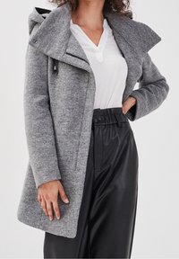 Manteau en tricot gris avec capuche, présentant un col asymétrique, des poches zippées et un tissu texturé. Associé à un pantalon noir plissé.