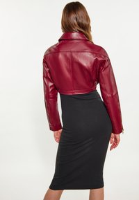 Giacca in pelle bordeaux tagliata, con un ampio colletto e maniche lunghe, abbinata a un vestito aderente nero. Texture liscia, senza motivi visibili.