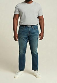 Homme portant un t-shirt gris clair, un jean bleu et des baskets blanches, debout devant un fond clair uni.