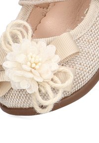 Zapato de tela beige con un diseño tejido texturizado. Presenta un acento de flor, cinta color crema y un detalle trenzado cerca del dedo del pie. Forma redondeada.
