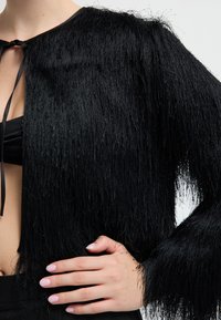 Czarna, teksturowana kurtka z frędzlami noszona na czarnym topie, z jedną ręką opartą na biodrze, widoczne jasnoróżowe paznokcie.