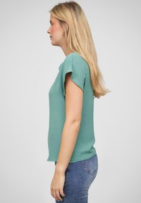 Lysegrønn kortermet topp med teksturert overflate, med en avslappet passform og subtile mansjetter, kombinert med blå denimjeans.