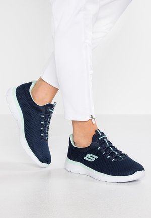 Personne portant des baskets Skechers bleu marine avec des accents vert clair et des semelles blanches, associées à un pantalon blanc avec des fermetures éclair aux chevilles.
