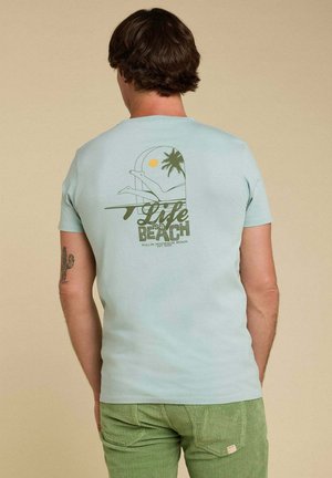T-shirt en coton bleu clair avec un graphique au dos représentant un palmier, un soleil et le texte "La vie est une plage" en vert et noir. Tissu doux et manches courtes.