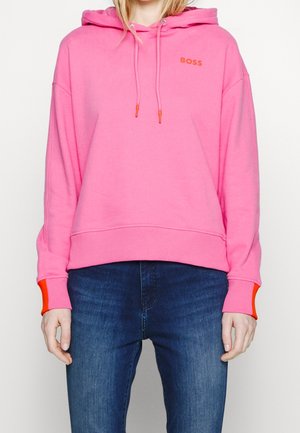 Personne portant un sweat à capuche rose avec un logo "BOSS" orange et des détails orange aux poignets, associée à un jean bleu, debout devant un fond uni.