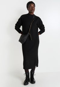 Robe tricot noire à manches longues avec un col côtelé et une fente sur le côté, associée à des bottes de combat noires à lacets et à un petit sac bandoulière noir.
