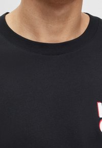 Close-up van een persoon die een zwart T-shirt met ronde hals draagt, met een gedeeltelijk rood en wit grafisch ontwerp op de onderrechte hoek.