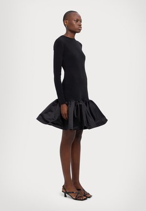 Robe noire à manches longues avec un buste ajusté et une jupe évasée en satin à volants. Portée avec des talons aiguilles noirs sur un fond neutre.