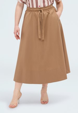 Mujer con falda midi beige con cintura de cordón, camiseta de manga corta a rayas y sandalias beige de tacón alto, de pie con una mano en el bolsillo.