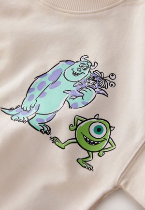 Sweatshirt en polaire beige avec des graphismes colorés de dessins animés montrant un monstre bleu à taches violettes et un monstre rond vert, tous deux souriants.