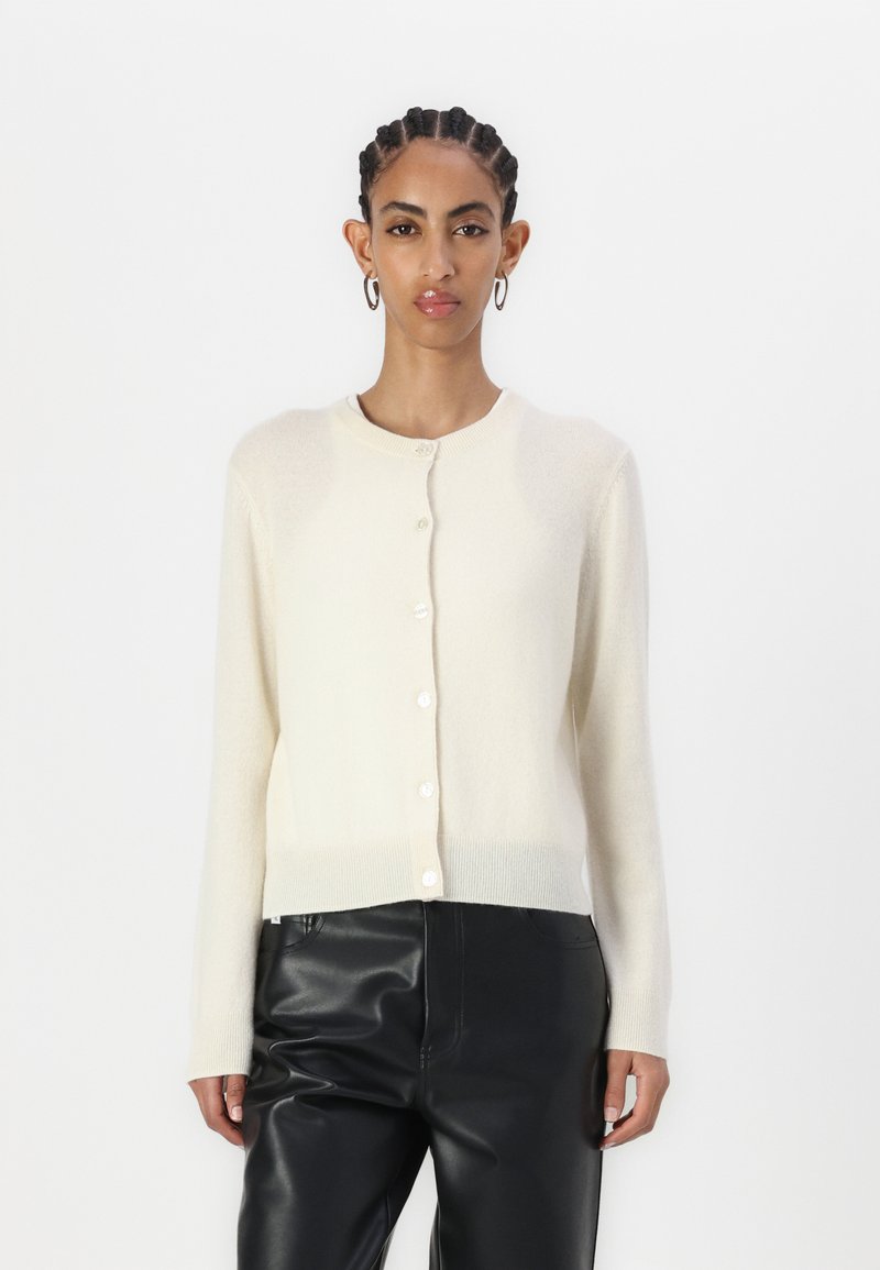 Cardigan couleur crème en matériau doux, avec un col rond, des manches longues et de petits boutons sur le devant. Porté avec un pantalon noir.