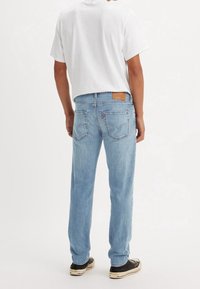 Personne portant un jean en denim bleu clair et un t-shirt blanc à manches courtes, debout de dos face au spectateur sur un fond blanc.