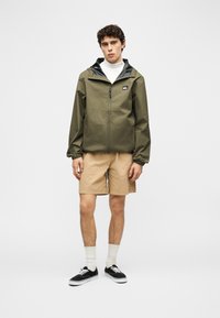 Νεαρός άνδρας στέκεται φορώντας ένα μπουφάν με φερμουάρ σε ελιά πράσινο χρώμα, μπεζ σορτς, λευκές κάλτσες και μαύρα αθλητικά παπούτσια σε απλό λευκό φόντο.