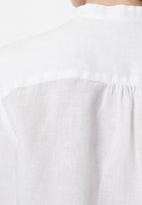 Chemise en lin blanc avec un empiècement froncé au dos et une texture douce et respirante. Le tissu présente un tissage léger et subtil.