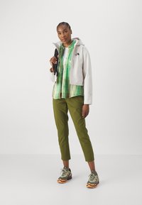 Hvit kort jakke med hette, grønn rutete skjorte, olivengrønne bukser og flerfargede sneakers. Svart ryggsekk holdt i den ene hånden.