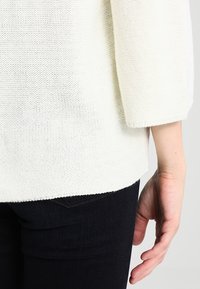 Persona che indossa un maglione bianco a maglia con maniche a tre quarti e pantaloni neri, mano rilassata al fianco.