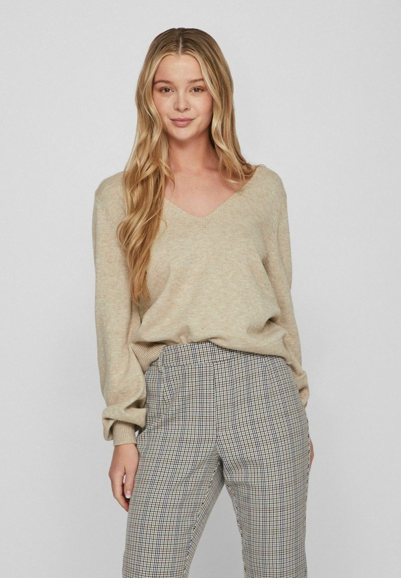 Beige gestrickter Pullover mit V-Ausschnitt, lockerem Schnitt und gerippten Bündchen, kombiniert mit karierten, taillierten Hosen in Schwarz und Weiß.