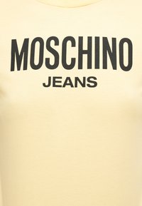 Ύφασμα σε κρεμ απόχρωση με μαύρο έντονο κείμενο "MOSCHINO" πάνω από μικρότερο "JEANS" στο κέντρο του.