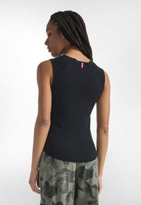 Mujer con cabello trenzado que lleva una blusa negra sin mangas y pantalones verdes con estampado floral, de espaldas contra un fondo blanco.