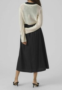 Žena stojící zády k fotoaparátu, oblečená v bílé síťované dlouhé tričko s dlouhým rukávem, černé midi sukni a černých lodičkách.