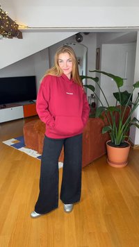 Maroon hoodie with front pocket and logo, paired with wide-leg black pants and silver shoes. Indoor setting with plants and furniture.