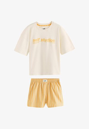 Persoon draagt lichtgele gestreepte korte broek met een witte tailleband met het label "self." en tilt een wit shirtje licht op.